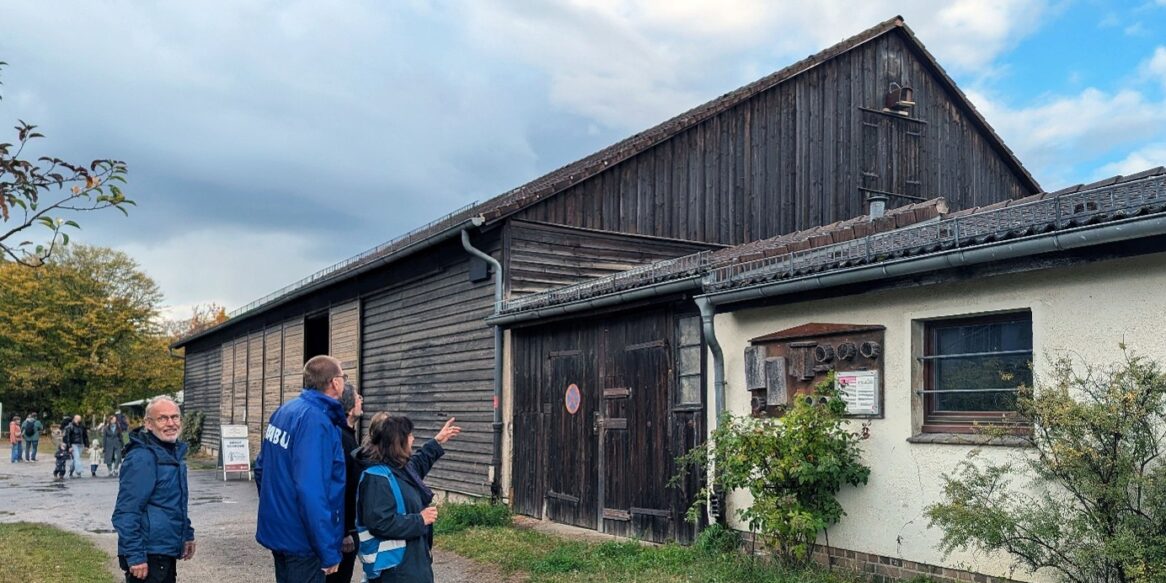 Fotografie einer historischen Scheune mit verwitterter Holzfassade, großem Tor, Ziegeldach und Anbau. Mehrere Mitglieder der NABU Bezirksgruppe Steglitz-Zehlendorf betrachten den ländlichen Gebäudekomplex unter bewölktem Himmel. Eine Frau zeigt auf den Turmfalken-Nistkasten, der am Giebel der Scheune angebracht wurde.