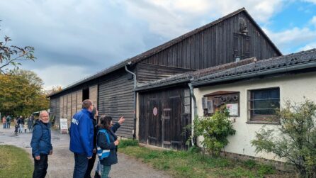 Fotografie einer historischen Scheune mit verwitterter Holzfassade, großem Tor, Ziegeldach und Anbau. Mehrere Mitglieder der NABU Bezirksgruppe Steglitz-Zehlendorf betrachten den ländlichen Gebäudekomplex unter bewölktem Himmel. Eine Frau zeigt auf den Turmfalken-Nistkasten, der am Giebel der Scheune angebracht wurde.